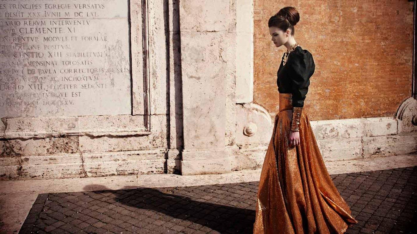 A model in an ochre pleated gown against a Roman stone facade