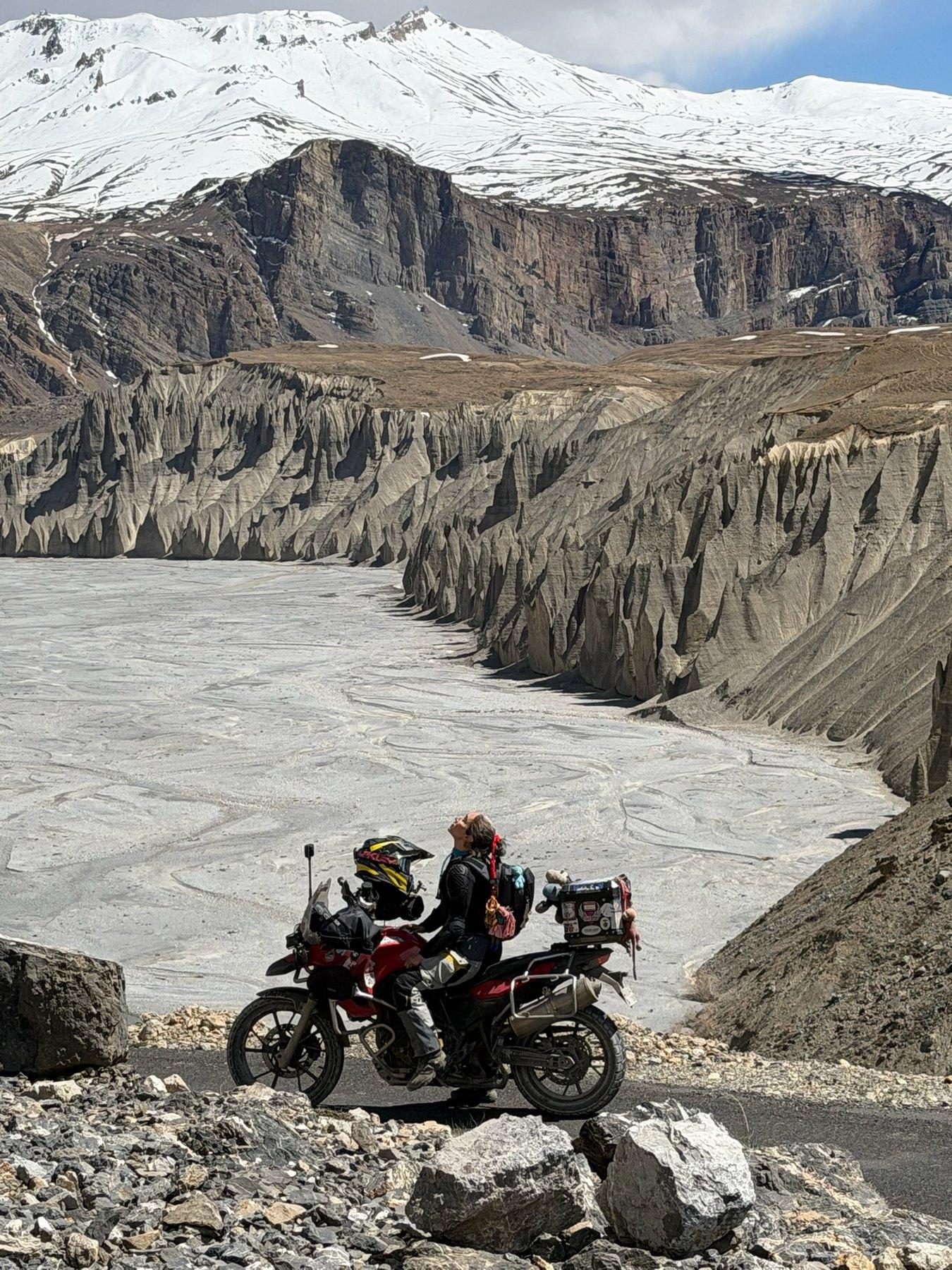 A loaded motorcycle on a mountain road above a vast river canyon, snow peaks behind